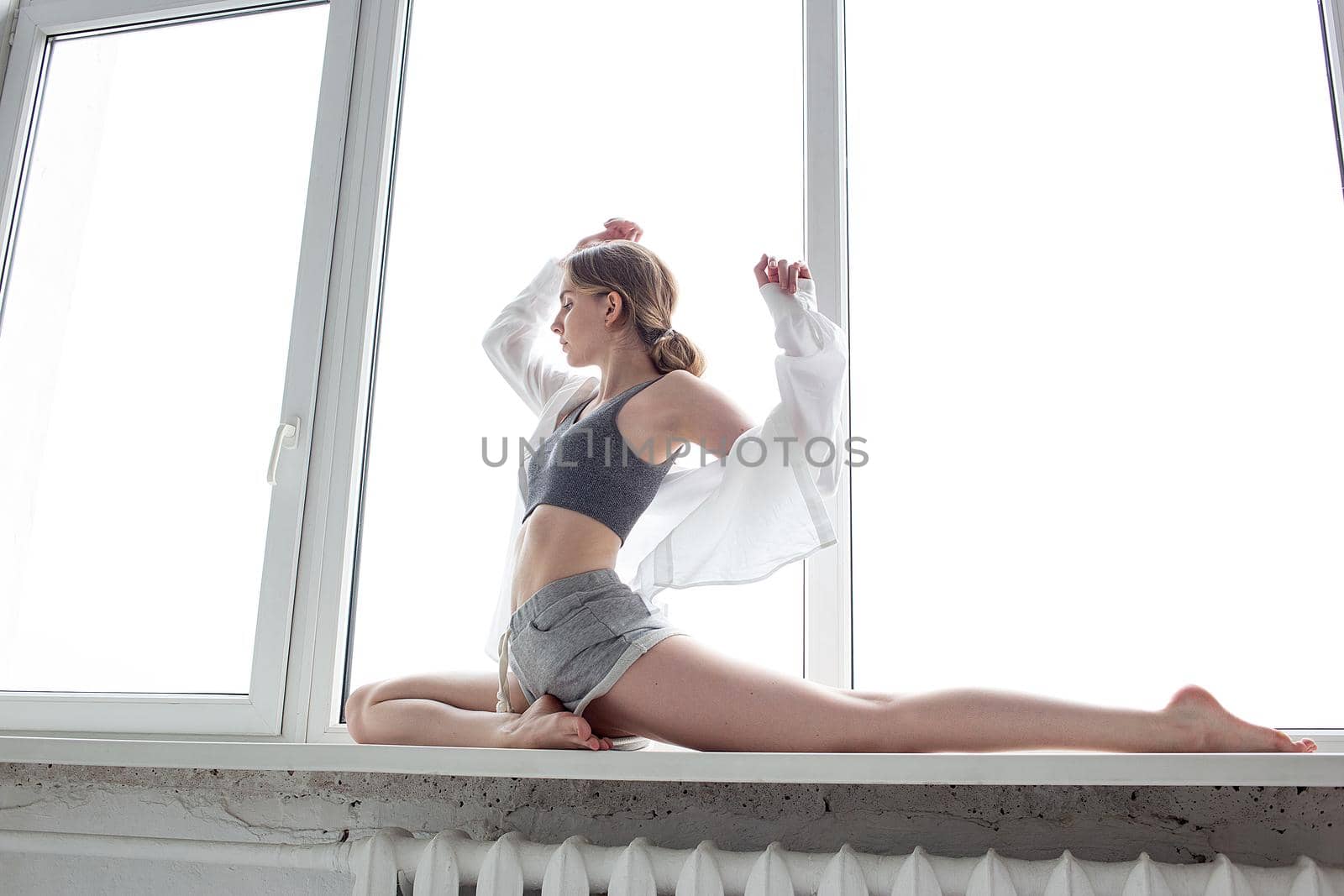 pretty girl in sportswear practicing yoga, doing splits, stretching exercise on windowsill at home. young attractive female with beautiful athletic body sitting on the splits, working out indoors