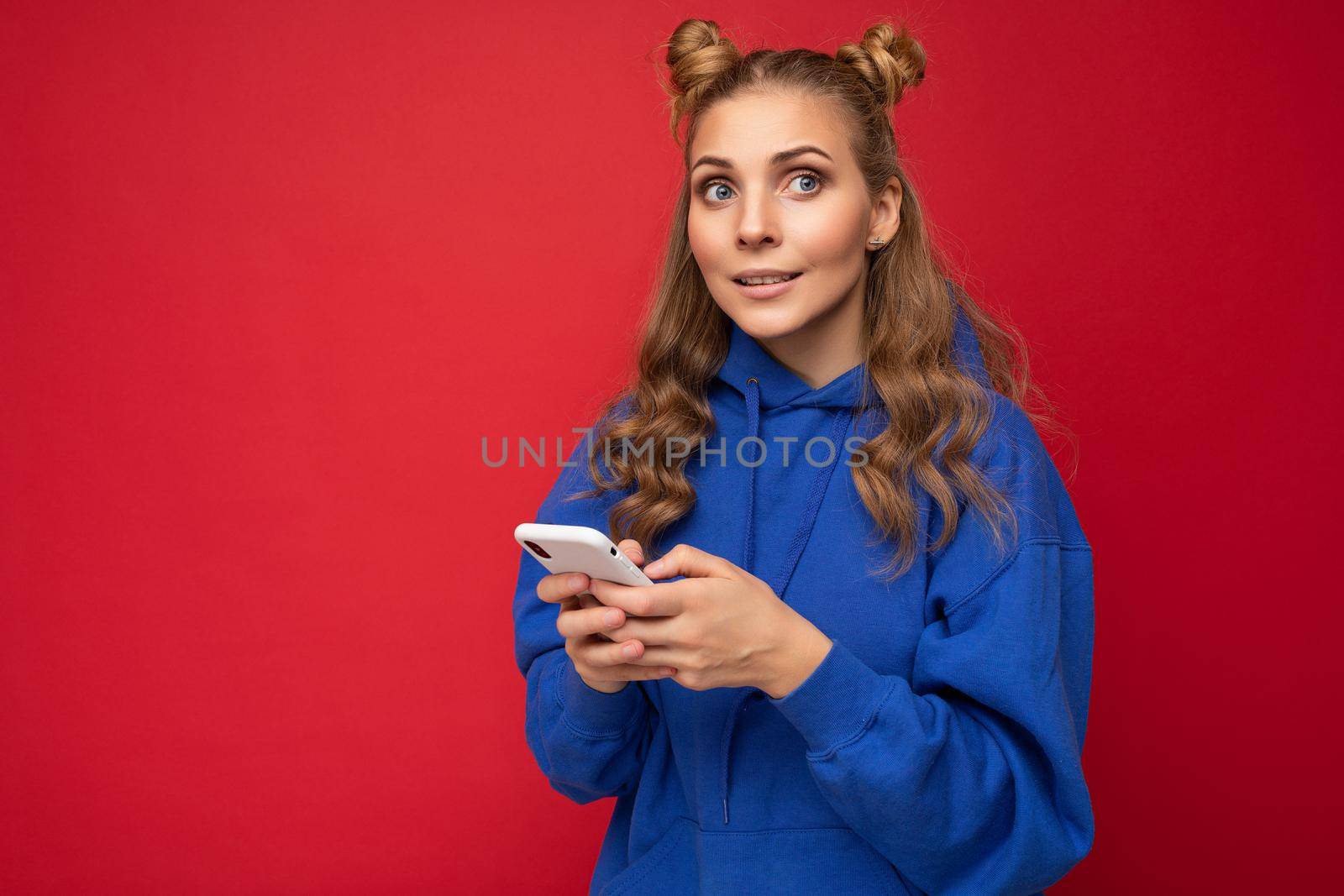 Attractive young blonde woman wearing blue stylish hoodie standing isolated over red background texting message via phone looking to the side.