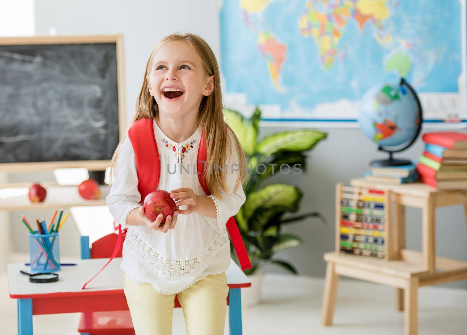 Little laughing blond girl standing near white desk with her red bag holding an apple in the spacious school classroom