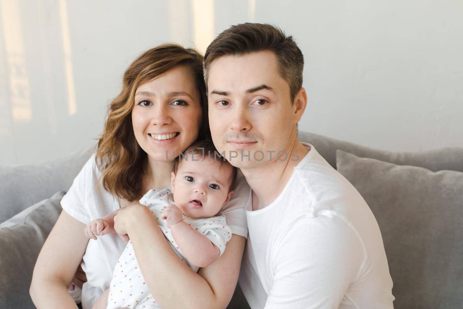 Happy young man and woman with newborn baby sitting on couch at home looking at camera