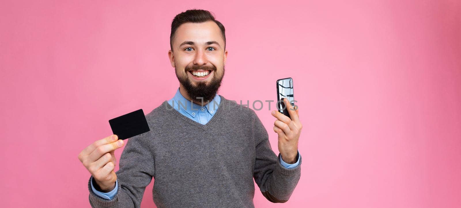 Panoramic photo shot of Handsome smiling brunette bearded young man wearing stylish grey sweater and blue shirt isolated over background wall holding credit card and mobile phone looking at camera.
