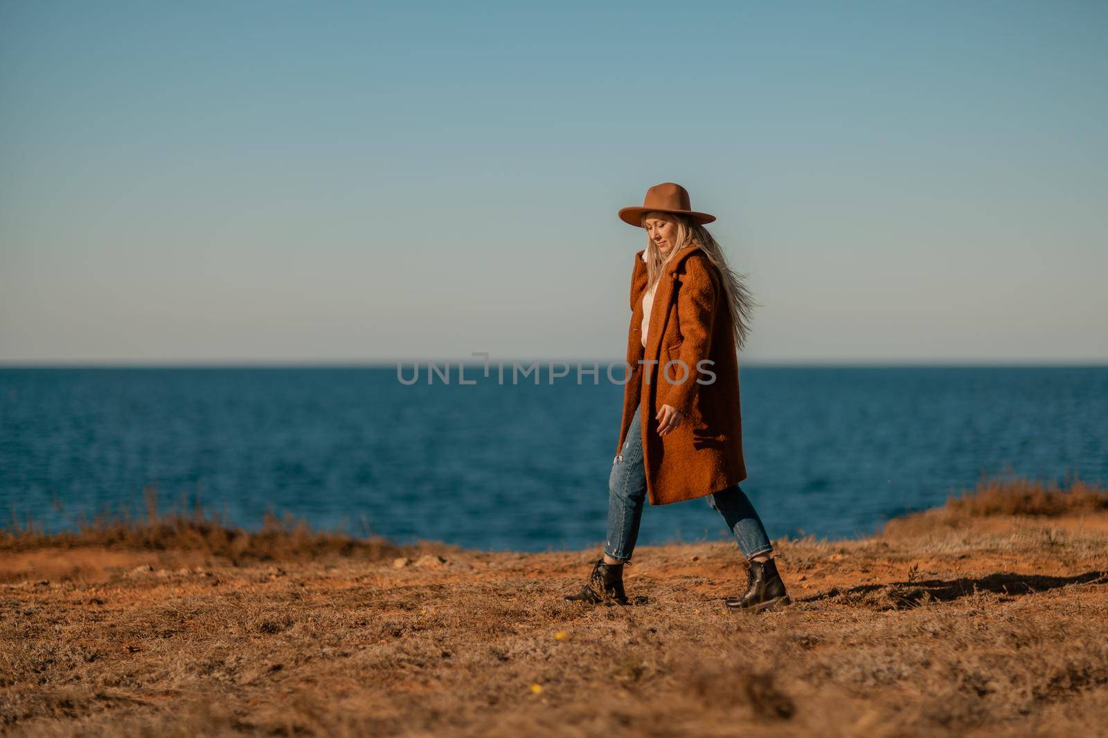 A woman walking along the coast near the sea. An elegant lady in a brown coat and a hat with fashionable makeup walks on the seashore.