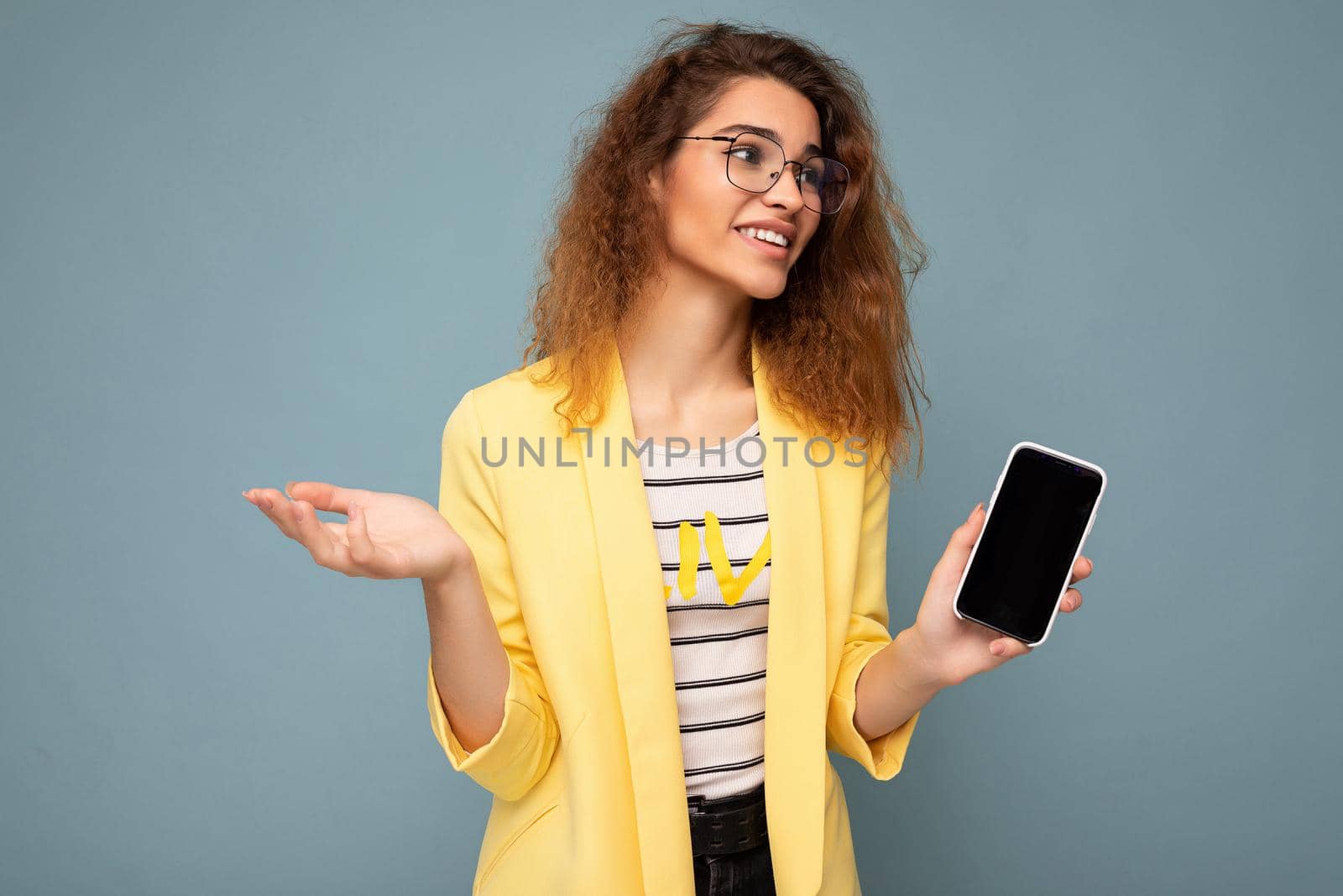 attractive young woman with curly dark blond hair wearing yellow jacket and optical glasses isolated on background holding and showing mobile phone with empty space for cutout looking to the side and asking.