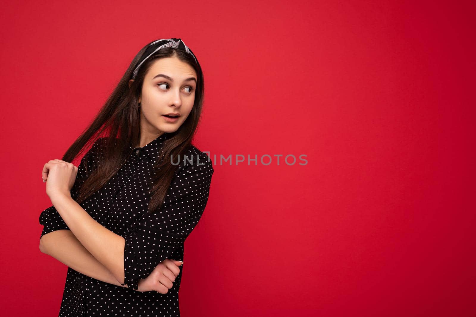 Photo of beautiful cute positive brunette girl standing isolated over red wall wearing casual stylish black clothes looking to the side. copy space