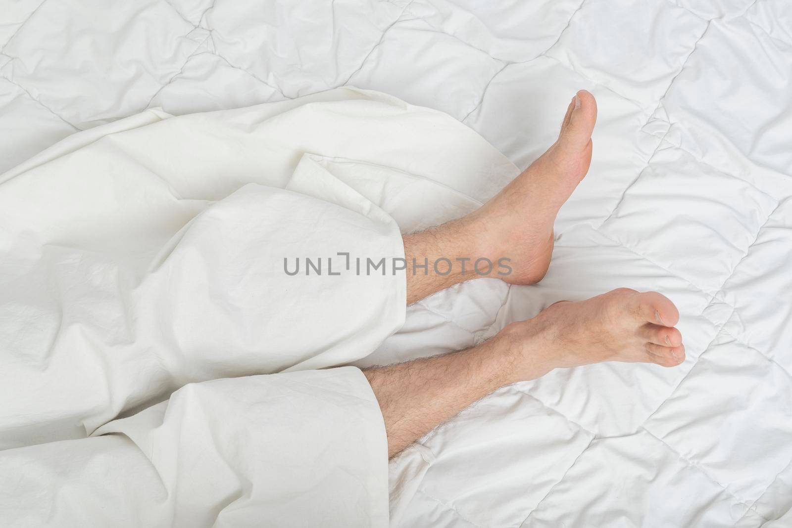 Photo of male feet on the bed under the sheet. Top view. Man sleeping on bed under blanket