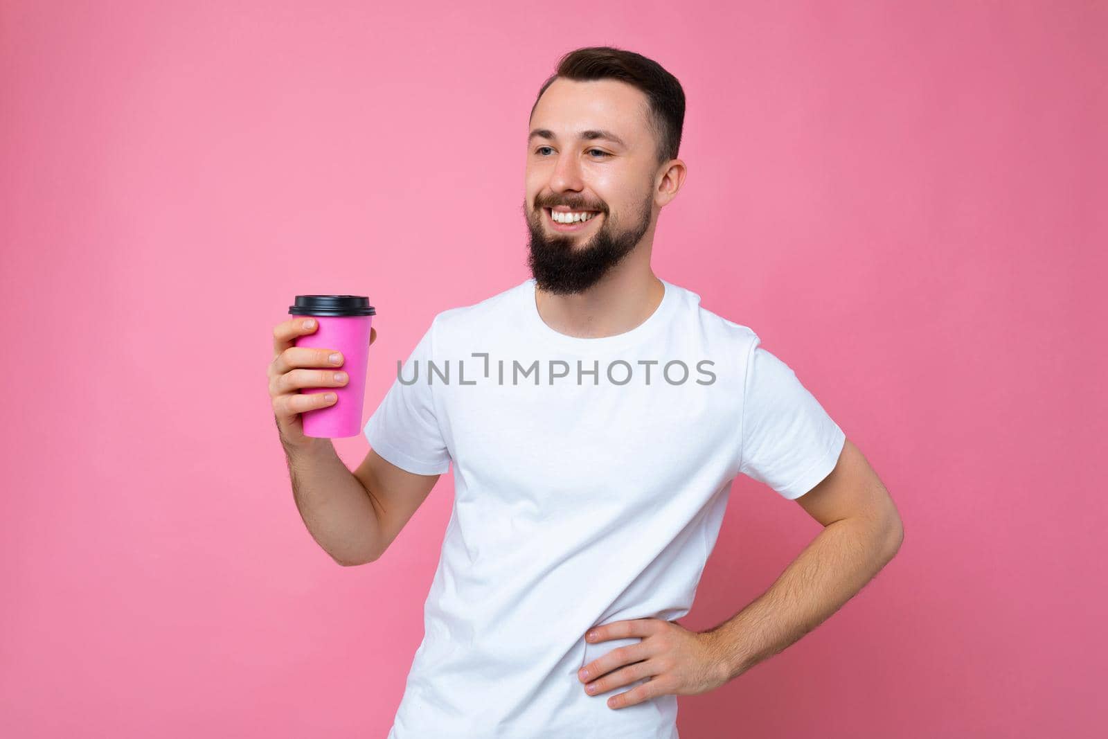 Handsome positive young brunette bearded man wearing white t-shirt isolated over pink backgroung wall holding paper coffee cup drinking and looking to the side.