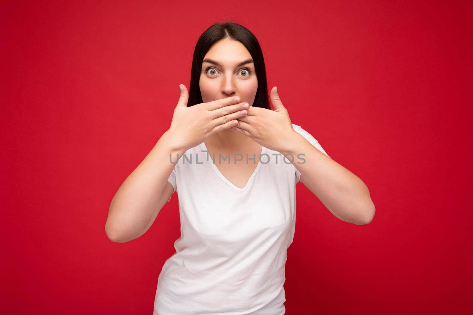 Portrait of young pretty nice brunette female person with sincere emotions wearing casual white t-shirt for mockup isolated over red background with copy space and covering mouth with hands by TRMK