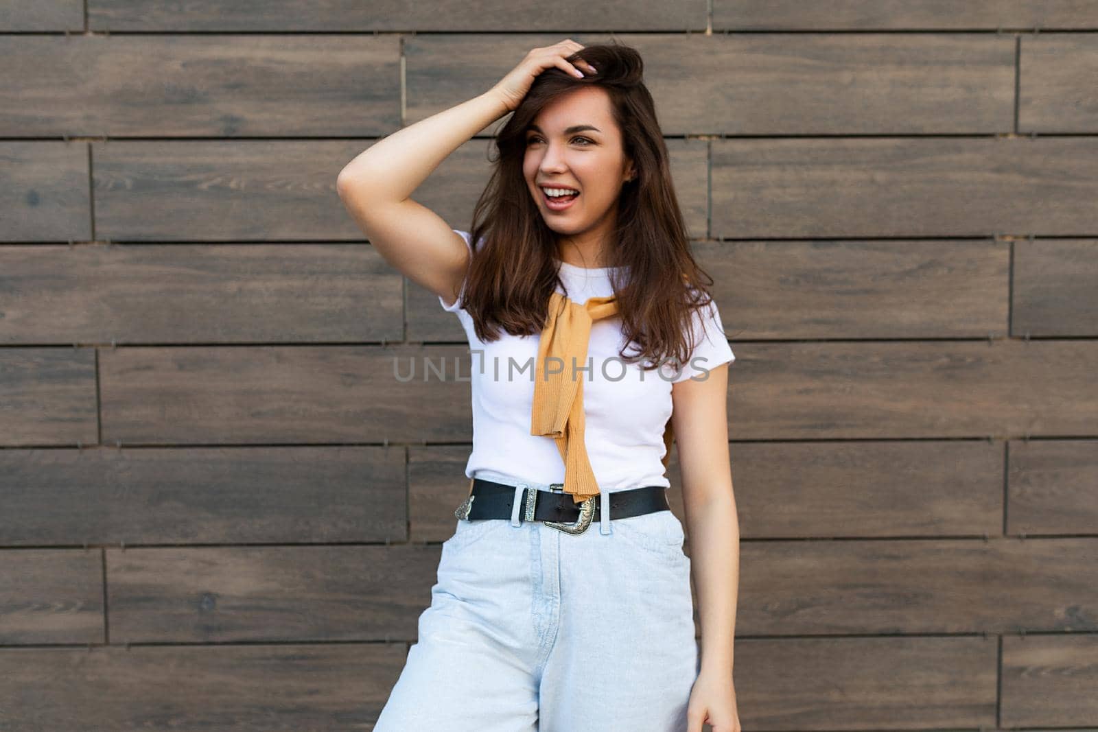 Portrait of successful smiling joyful happy young brunet woman wearing casual white t-shirt and jeans with yellow sweater poising near brown wall in the street and having fun.