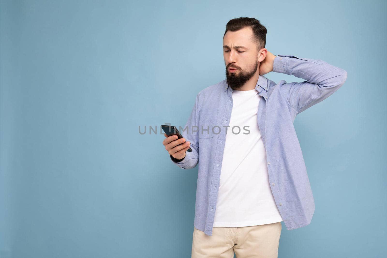 Photo shot of handsome thoughtful positive good looking young man wearing casual stylish outfit poising isolated on background with empty space holding in hand and using mobile phone messaging sms looking at smartphone display screen.