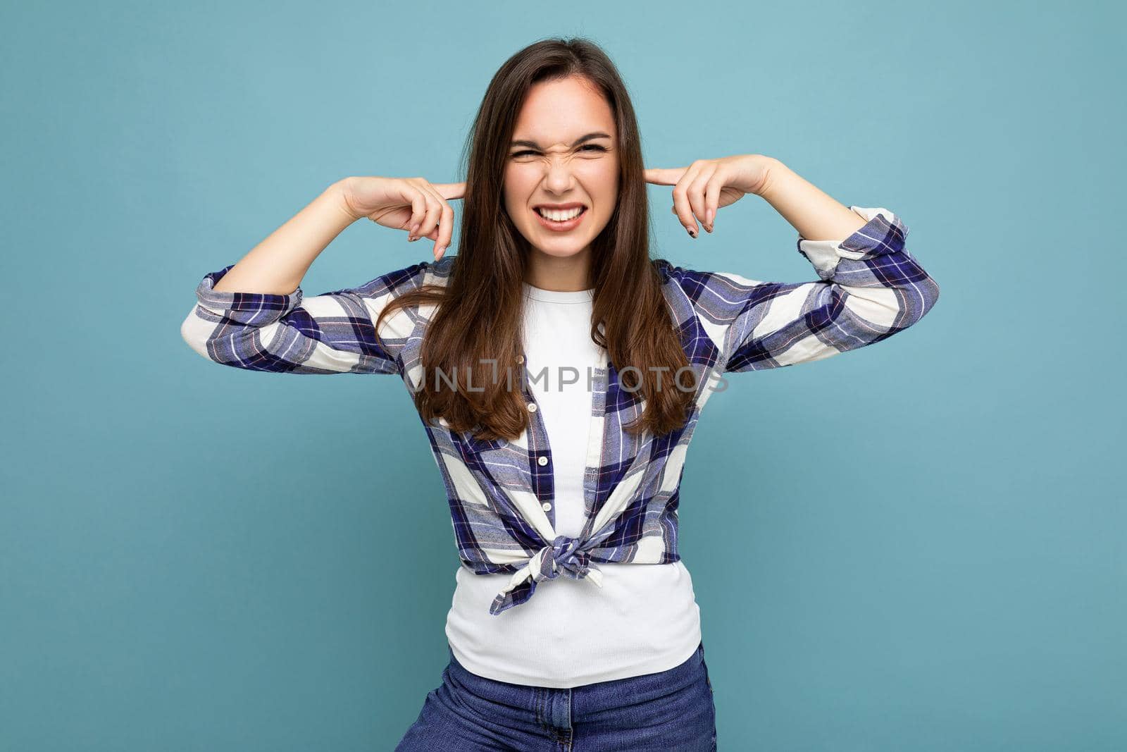 Don't want to hear it. Young emotional positive attractive brunette female person isolated on blue background with copy space and covering ears.