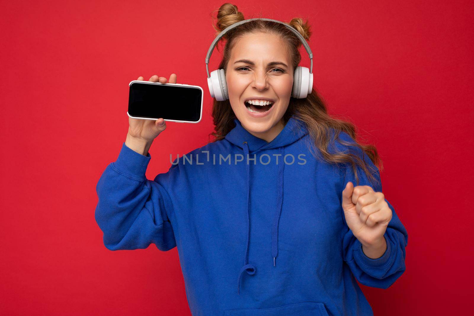 Beautiful happy smiling young woman wearing stylish casual outfit isolated on background wall holding and showing mobile phone with empty display for mockup wearing white bluetooth headphones listening to music and having fun looking at camera by TRMK