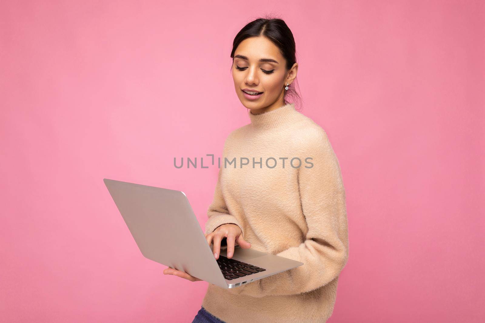 Close-up portrait of Beautiful young brunette woman wearing beige sweater holding netbook computer typing text on keyboard looking down isolated over pink wall background.