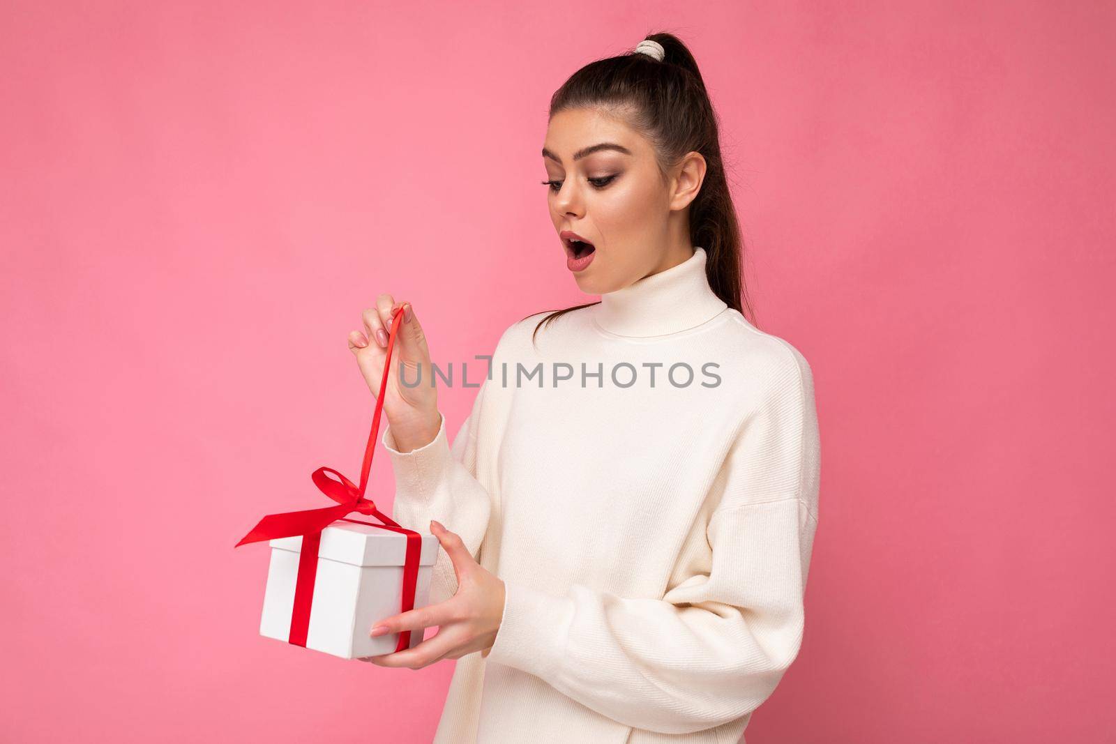 Photo shot of pretty positive surprised young brunet woman isolated over colourful background wall wearing trendy outfit look holding gift box and looking at present box with red ribbon by TRMK