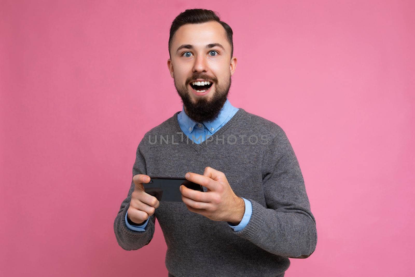 Shot of happy handsome young brunette unshaven man with beard wearing everyday grey sweater and blue shirt isolated on background wall holding smartphone playing games via mobile phone looking at camera and having fun by TRMK
