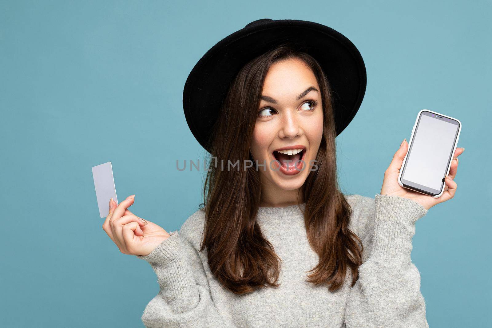 Closeup portrait photo of attractive joyful young brunette woman wearing black hat and grey sweater isolated over blue background holding credit card and mobile phone with empty display for mockup looking to the side.