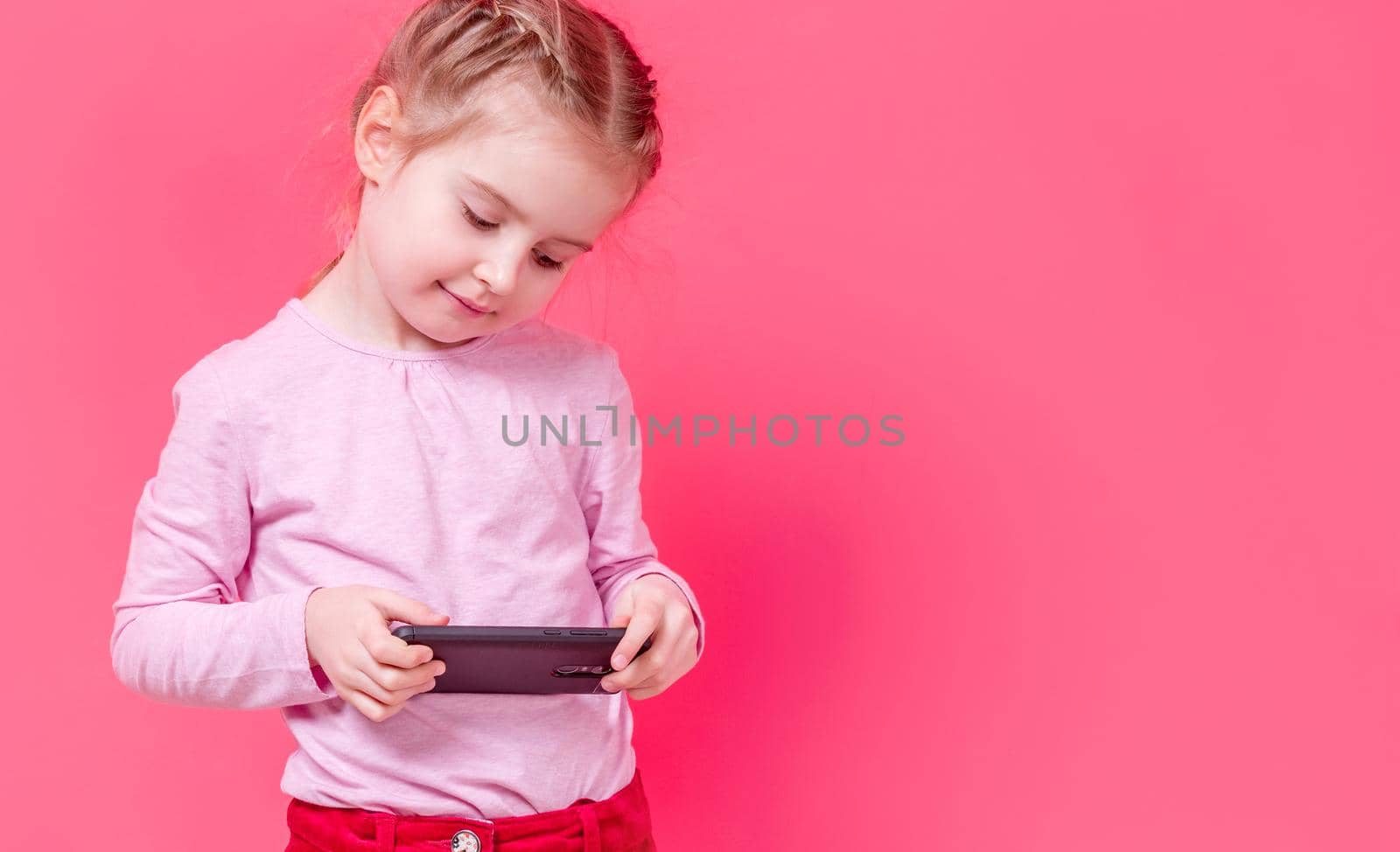 Adorable stylish small girl using smartphone over pink background