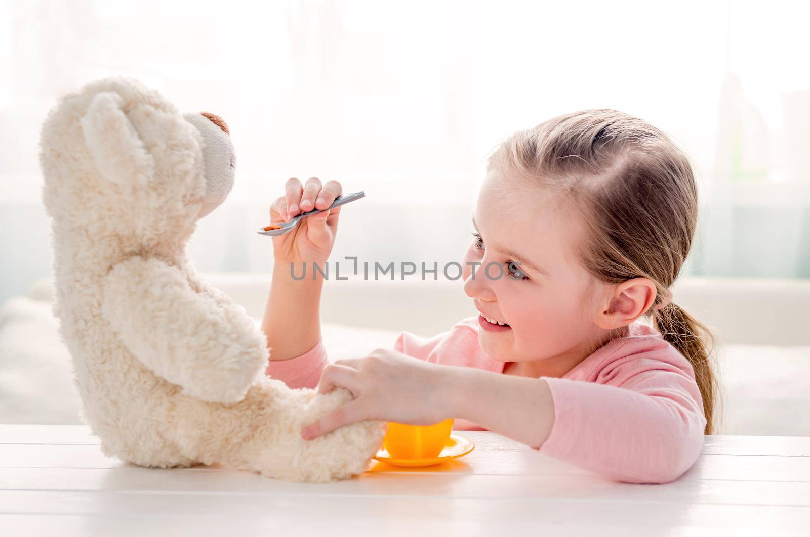 Cute little girl feeding her teddy bear with spoon. Beautiful child girl sitting at a table and feed teddy bear. Happy little kid playing with toy teddy bear