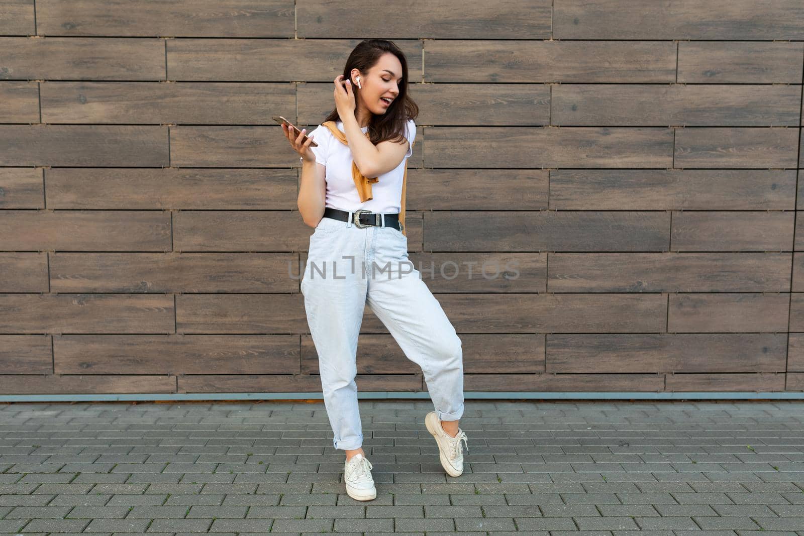 Full length body size photo of beautiful joyful smiling young woman wearing stylish casual clothes standing in the street holding and using mobile phone wearing white bluetooth headphones listening to music and having fun looking to the side.