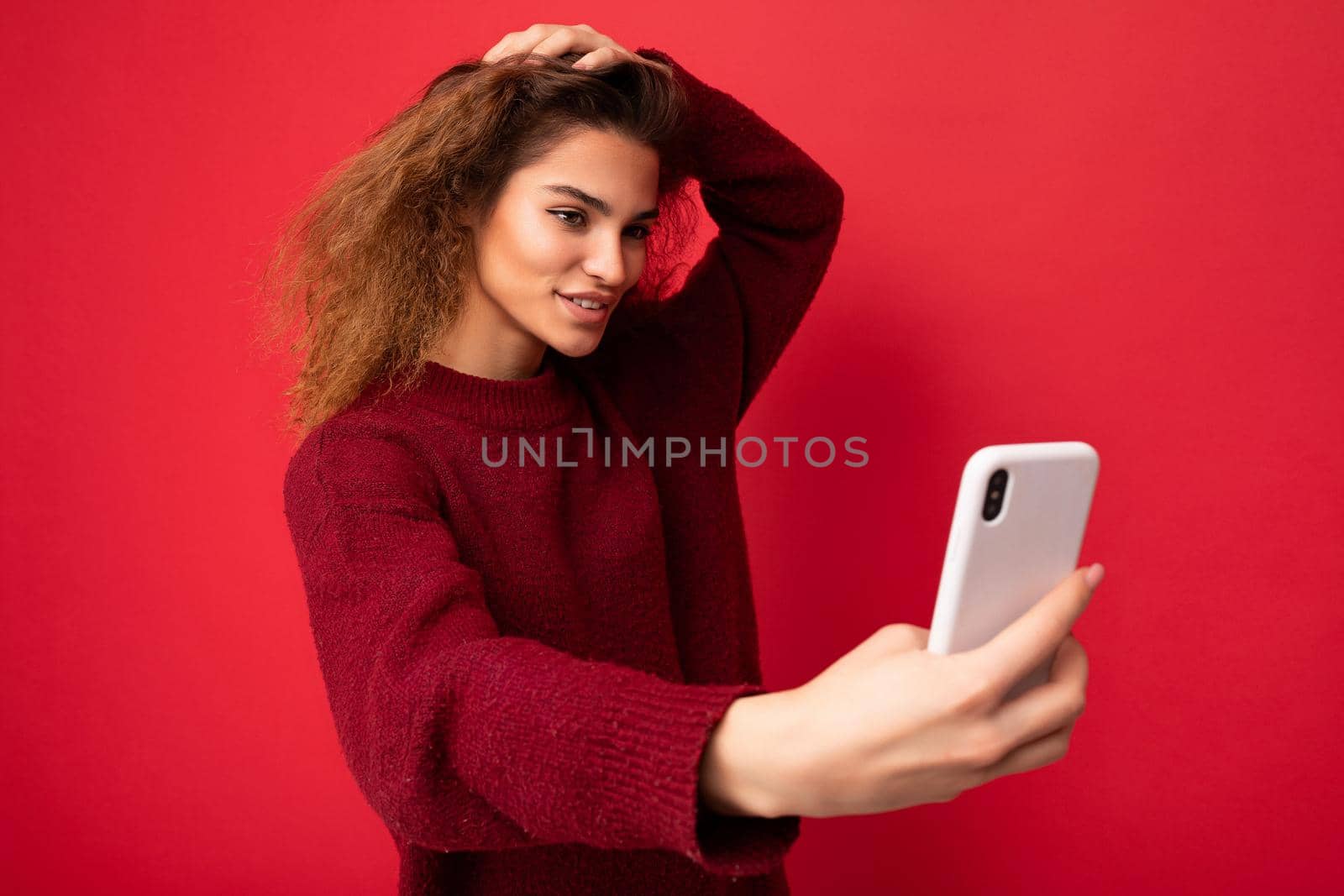 Shot of sexy smiling Beautiful young woman with curly hair wearing dark red sweater isolated on red background wall holding and using smart phone looking at telephone screen and taking selfie.