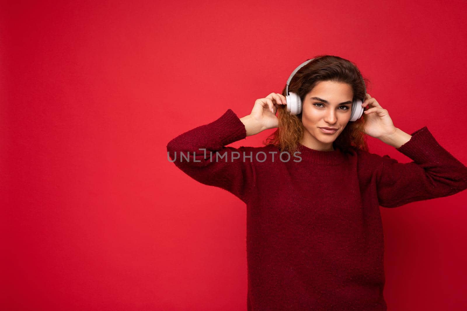 Beautiful happy smiling young brunette curly woman wearing dark red sweater isolated over red background wall wearing white bluetooth headphones listening to music and having fun looking at camera by TRMK