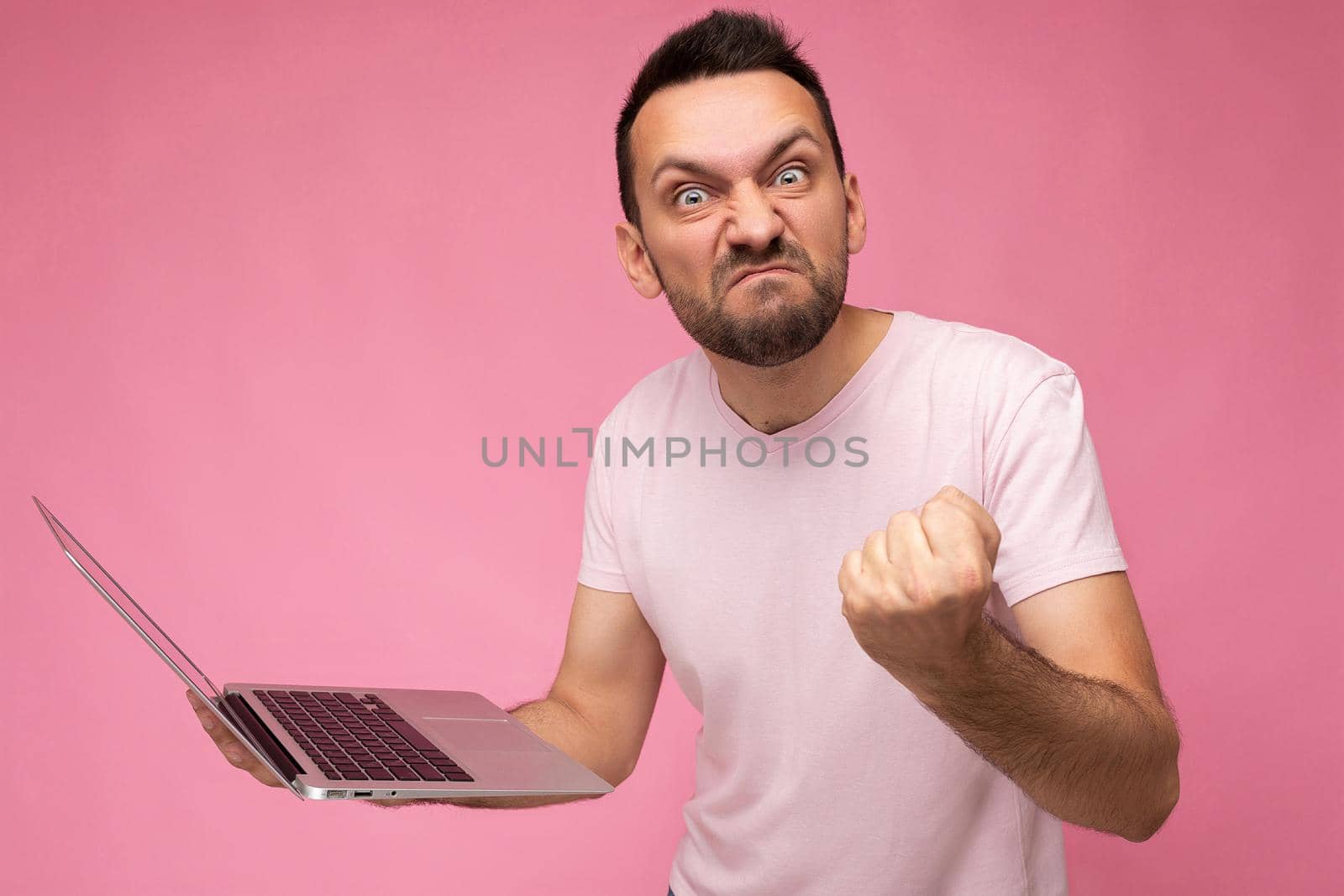 Handsome angry man holding laptop computer showing fist looking at camera in t-shirt on isolated pink background by TRMK