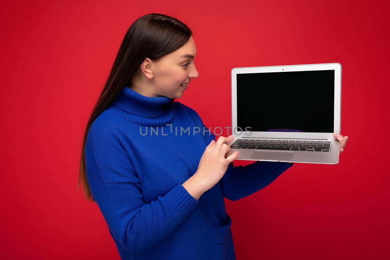 Photo shot of beautiful young brunet woman holding computer laptop looking at netbook keyboard in blue sweater isolated over red wall background. Cutout