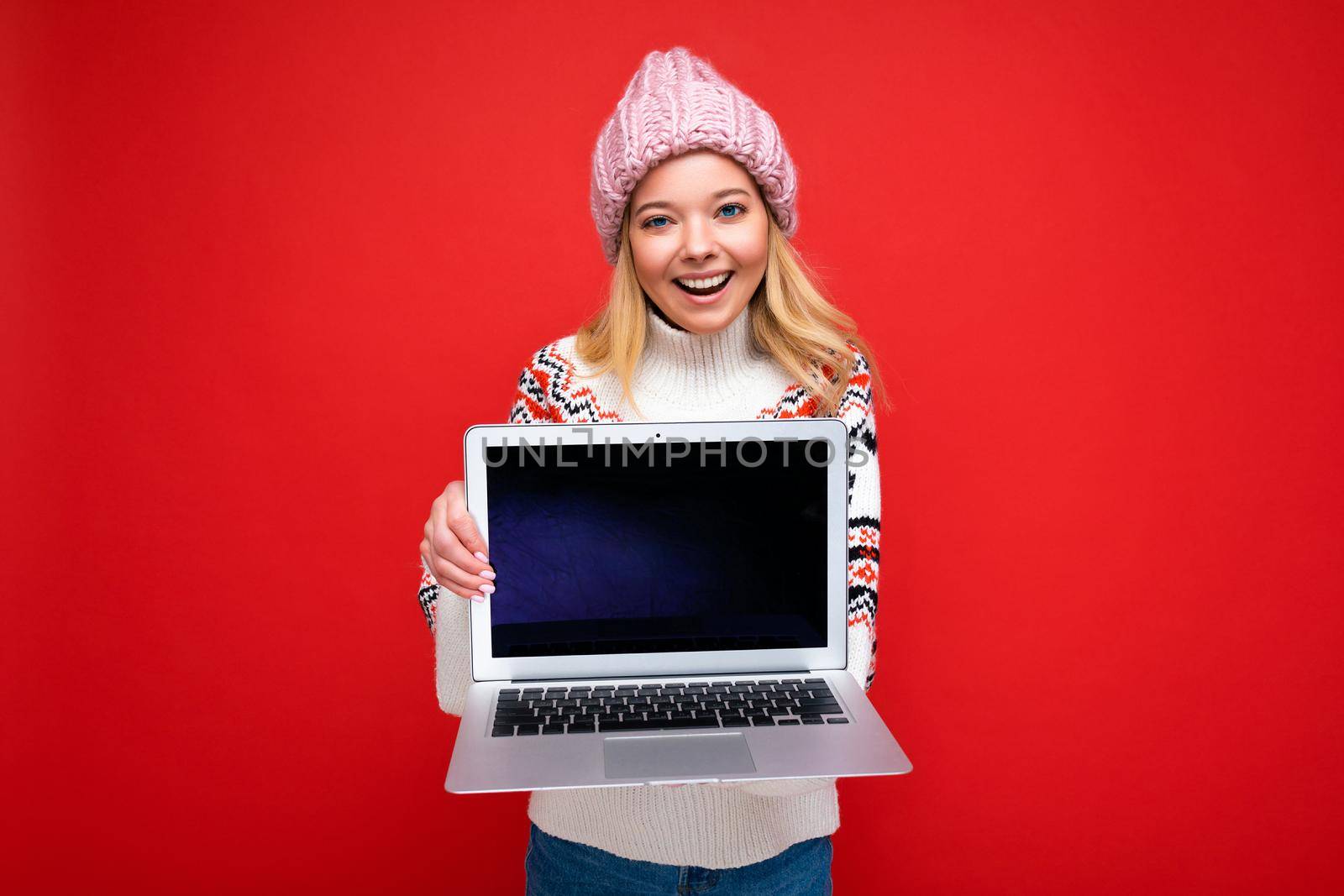 Photo portrait shot of beautiful smiling blonde young woman holding computer laptop with empty monitor screen with mock up and copy space wearing knitted winter hat and sweater looking at camera isolated over red wall background and winking.