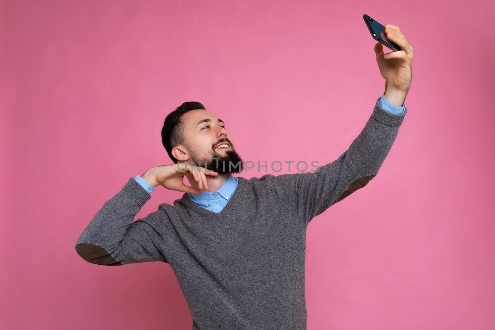 Photo of positive handsome young brunette unshaven man with beard wearing casual grey sweater and blue shirt isolated on pink background wall holding smartphone taking selfie photo looking at mobile phone screen display.