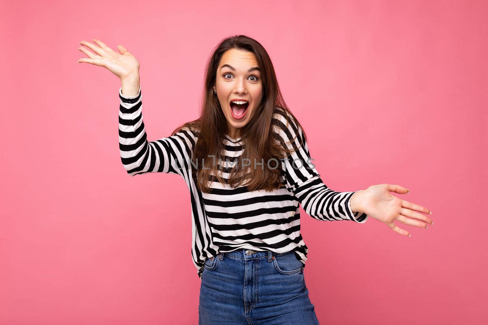 Photo of young emotional astonished shocked positive happy beautiful brunette woman with sincere emotions in casual striped pullover isolated on pink background with copy space.