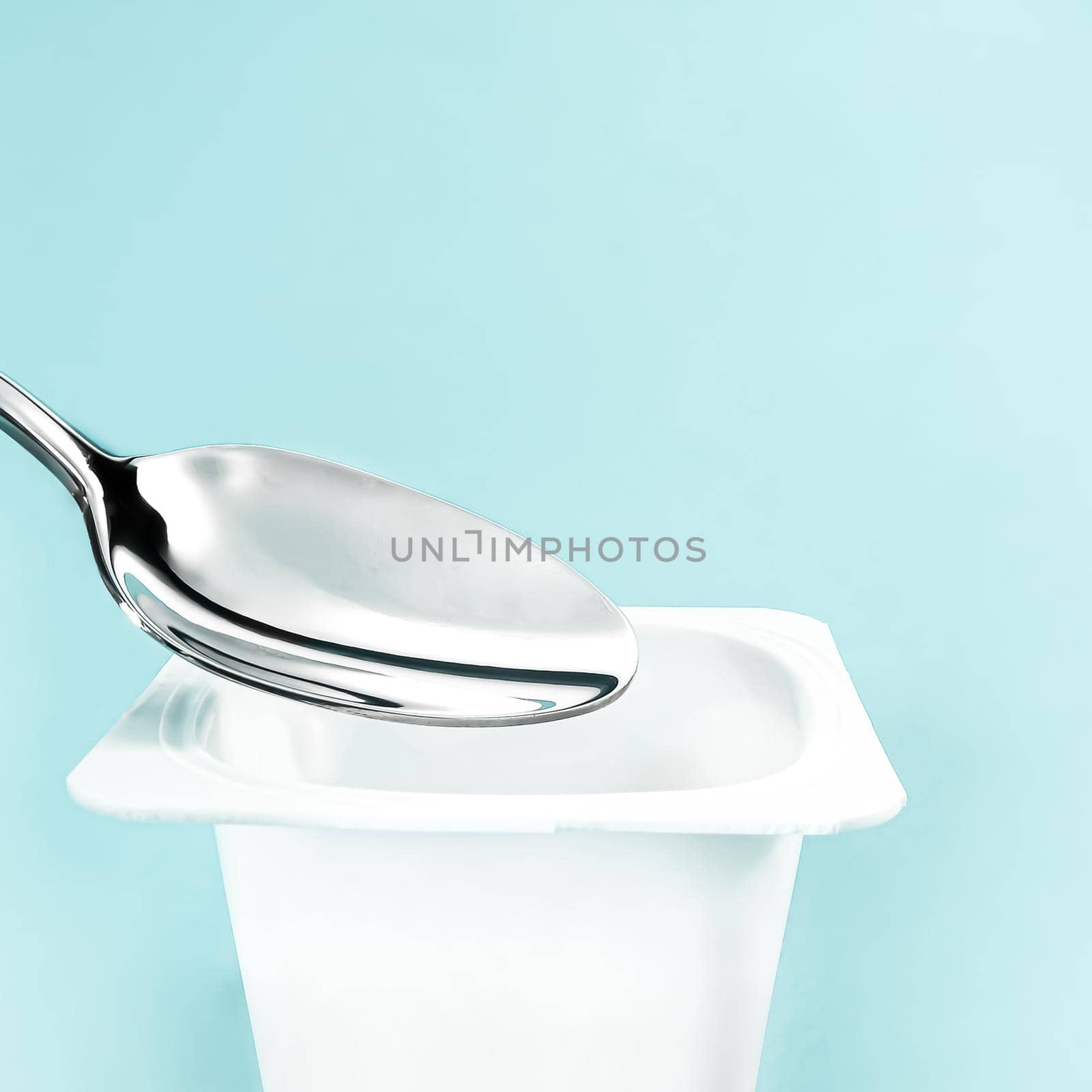 Yogurt cup and silver spoon on mint background, white plastic container with yoghurt cream, fresh dairy product for healthy diet and nutrition balance.