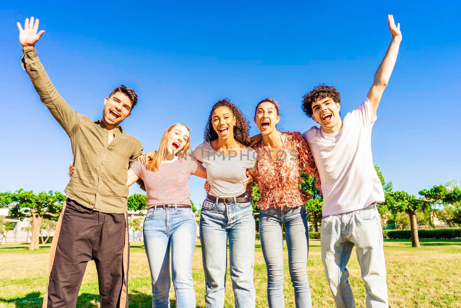 Multiracial group of young friends posing with raised open arms looking at camera embracing to each other in nature of city park. Happy diverse people having fun together enjoying life and success