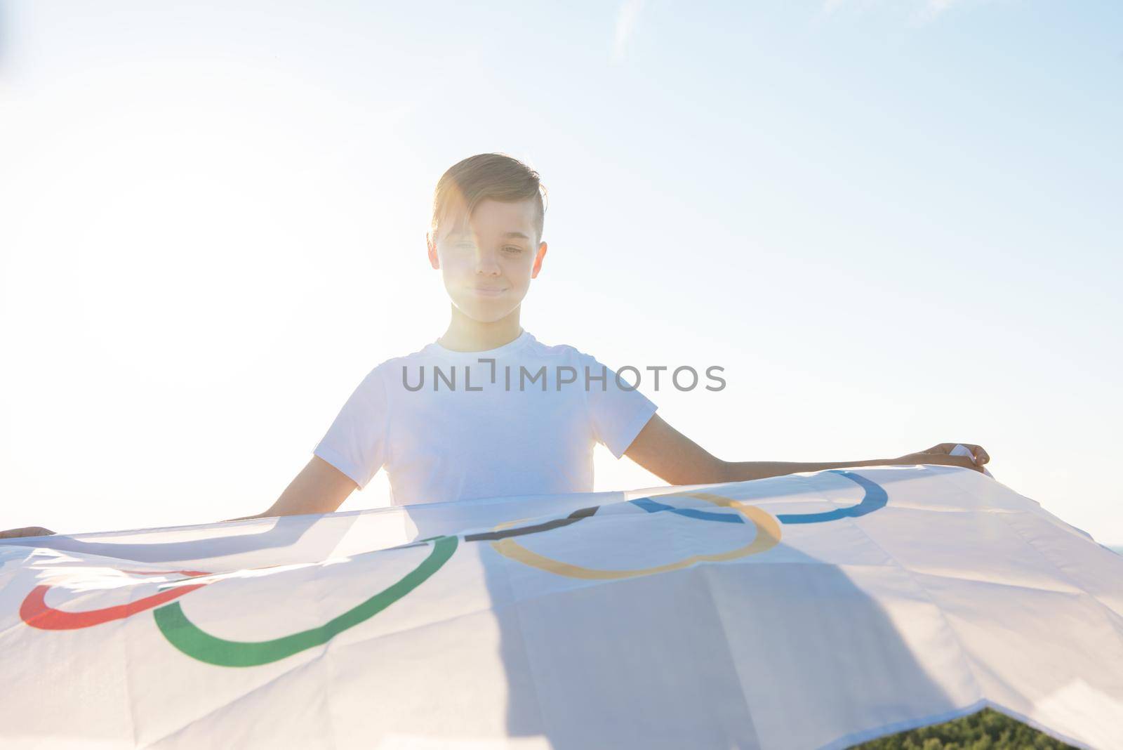Blonde boy waving waving flag the Olympic Games outdoors over blue sky at the river bank by rusak