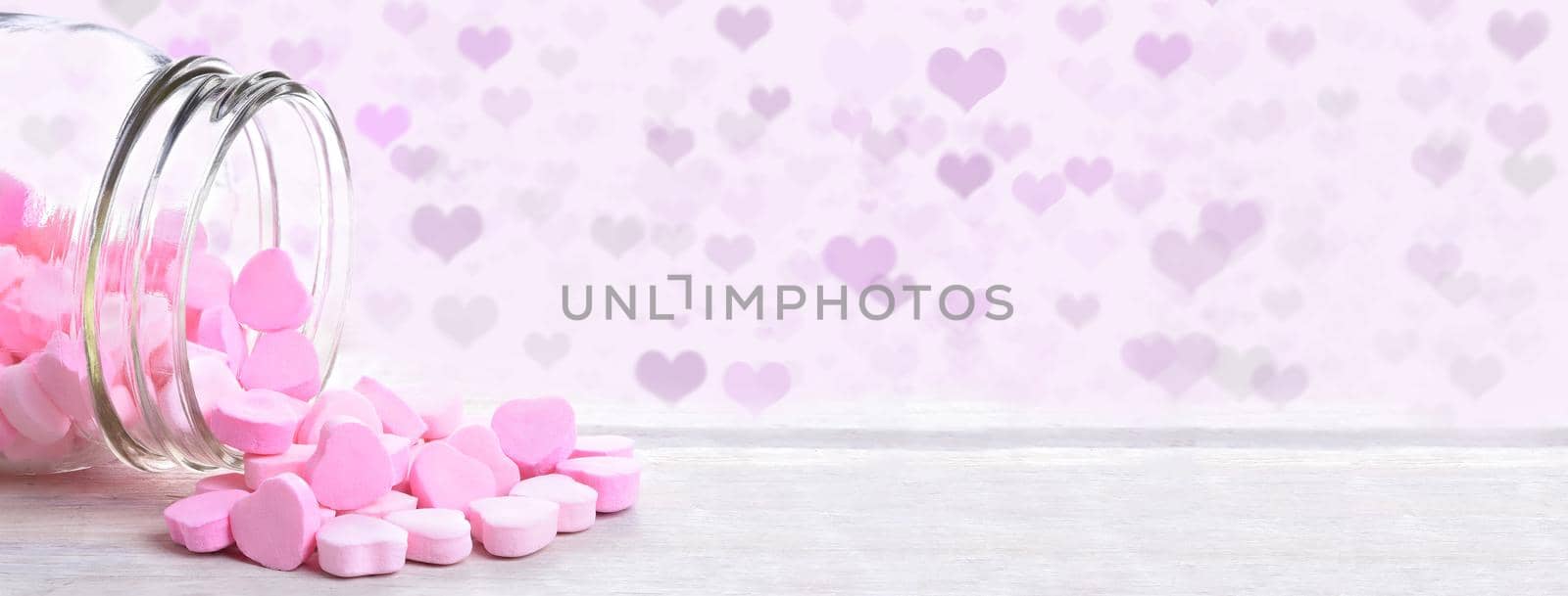 A jar of valentines heart shaped candy spilling onto a rustic wood table with heart Bokeh background.