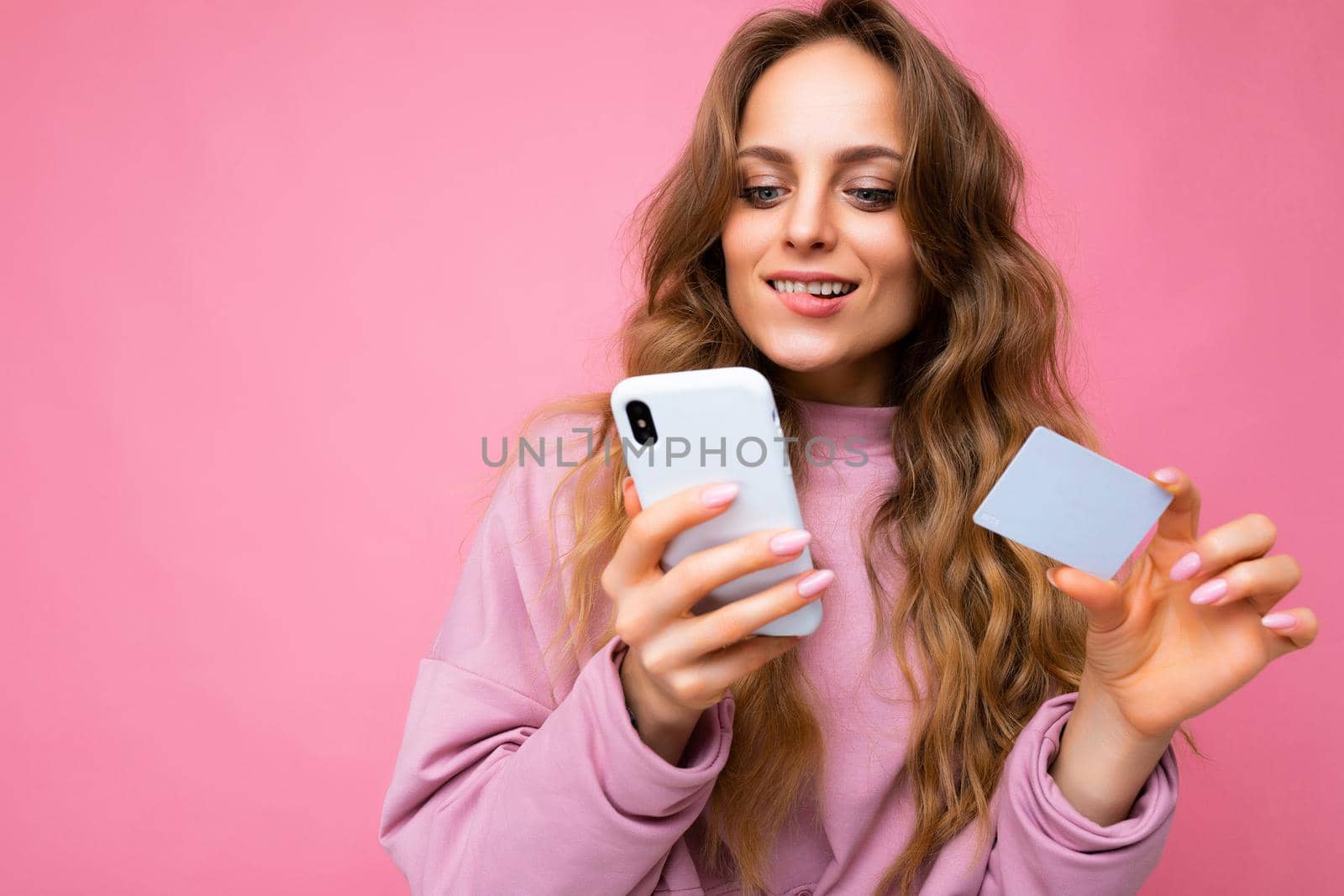 Beautiful sexy positive young blonde curly woman wearing pink clothes isolated over pink background using mobile phone paying online shopping through credit card looking at smartphone display.