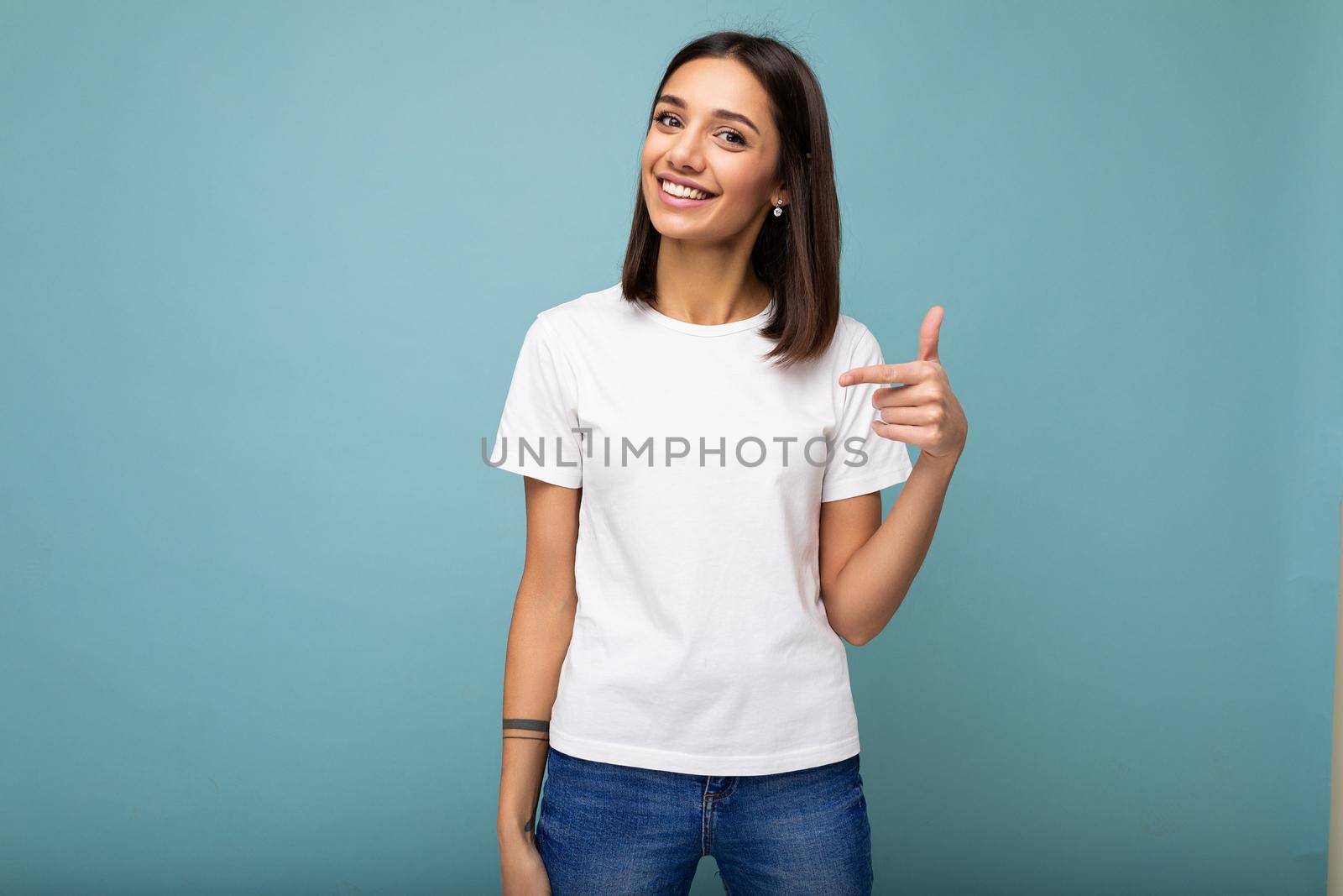 Portrait of young beautiful happy smiling brunette woman wearing trendy white t-shirt with empty space for mock up. Sexy carefree female person posing isolated near blue wall in studio with free space. Positive model with natural makeup by TRMK
