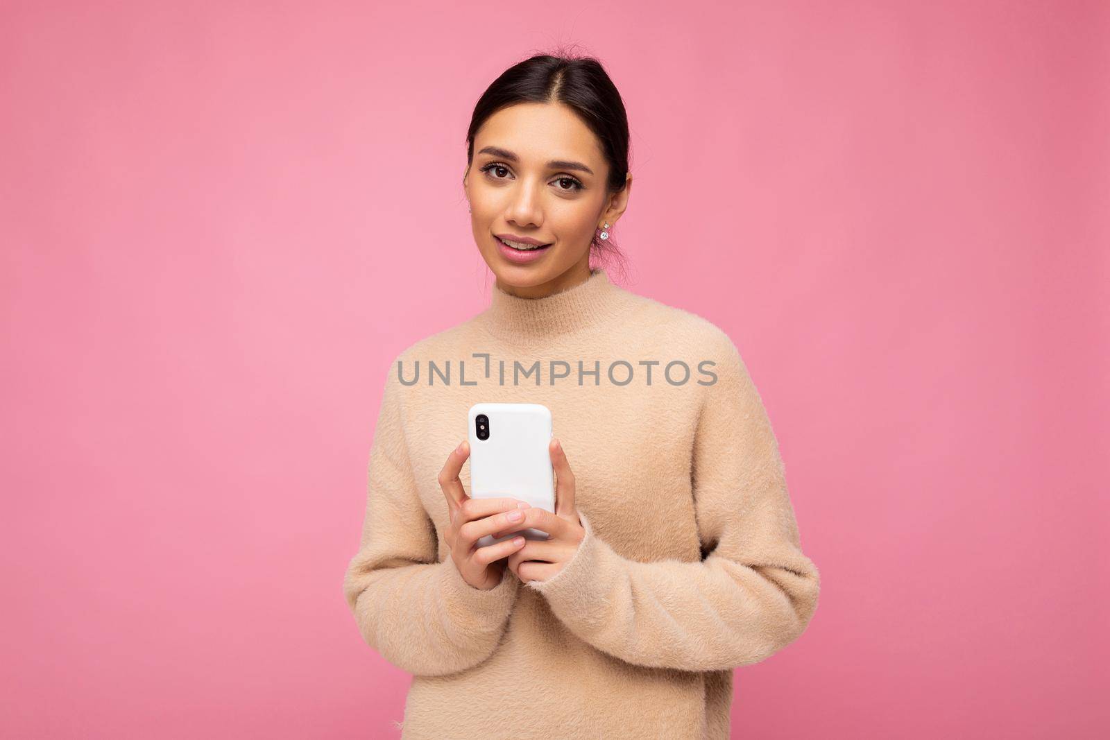 Attractive cute young brunet woman wearing beige warm sweater standing isolated over pink background surfing on the internet via phone looking at camera.