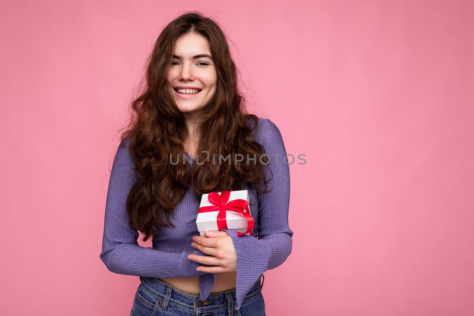 Beautiful happy young brunette woman isolated over colourful background wall wearing stylish casual clothes holding gift box and looking at camera by TRMK