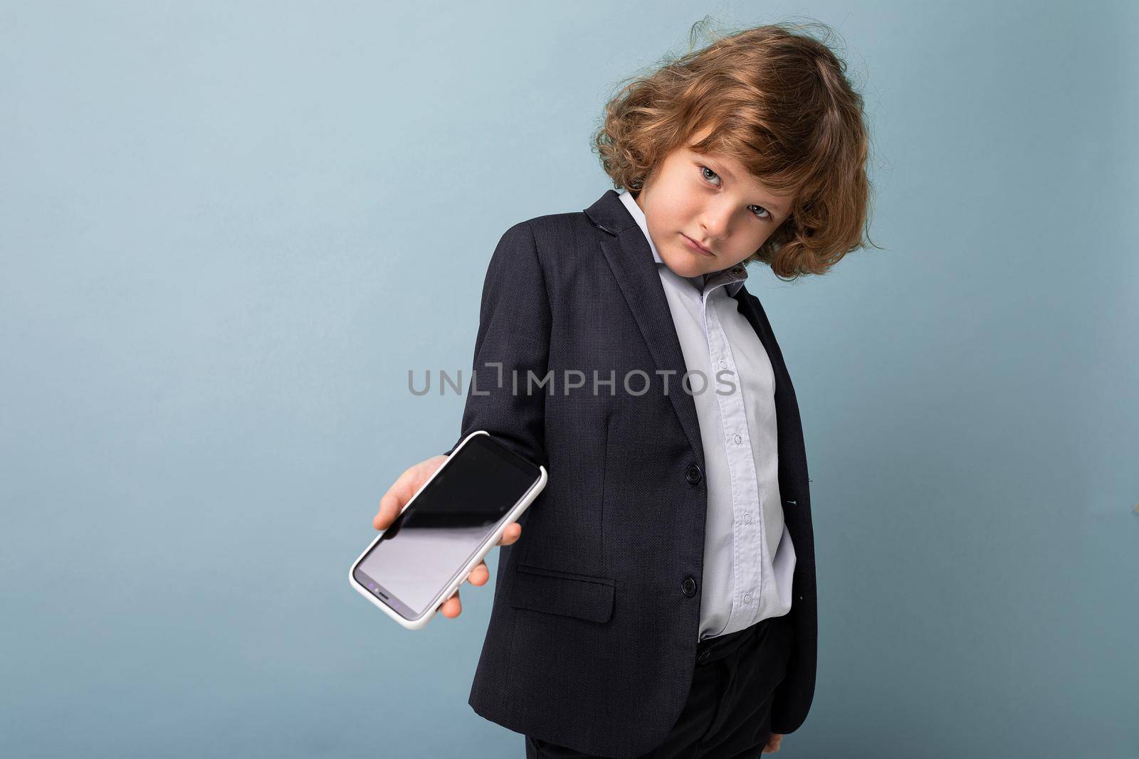 Handsome positive boy with curly hair wearing suit holding phone isolated over blue background looking at camera and showing smartphone with empty display screen by TRMK