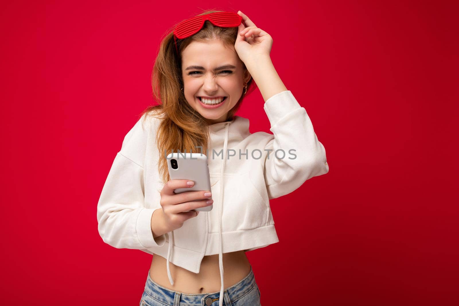 Beautiful emotional positive young blonde woman wearing stylish white hoodie and funny colorful glasses standing isolated over colourful background holding and using mobile phone communicating online on the internet by TRMK