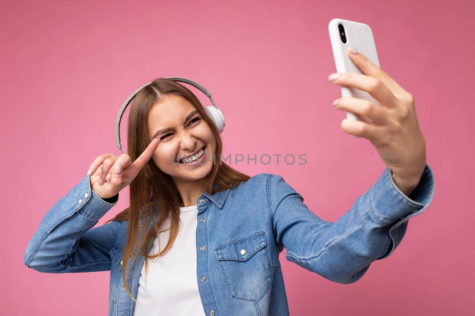 Photo of attractive happy smiling young blonde female person wearing blue jean stylish shirt and casual white t-shirt isolated over pink background wearing white bluetooth wireless headphones and listening to good music and using mobile phone taking selfie photo looking at gadjet and pointing finger at device camera.