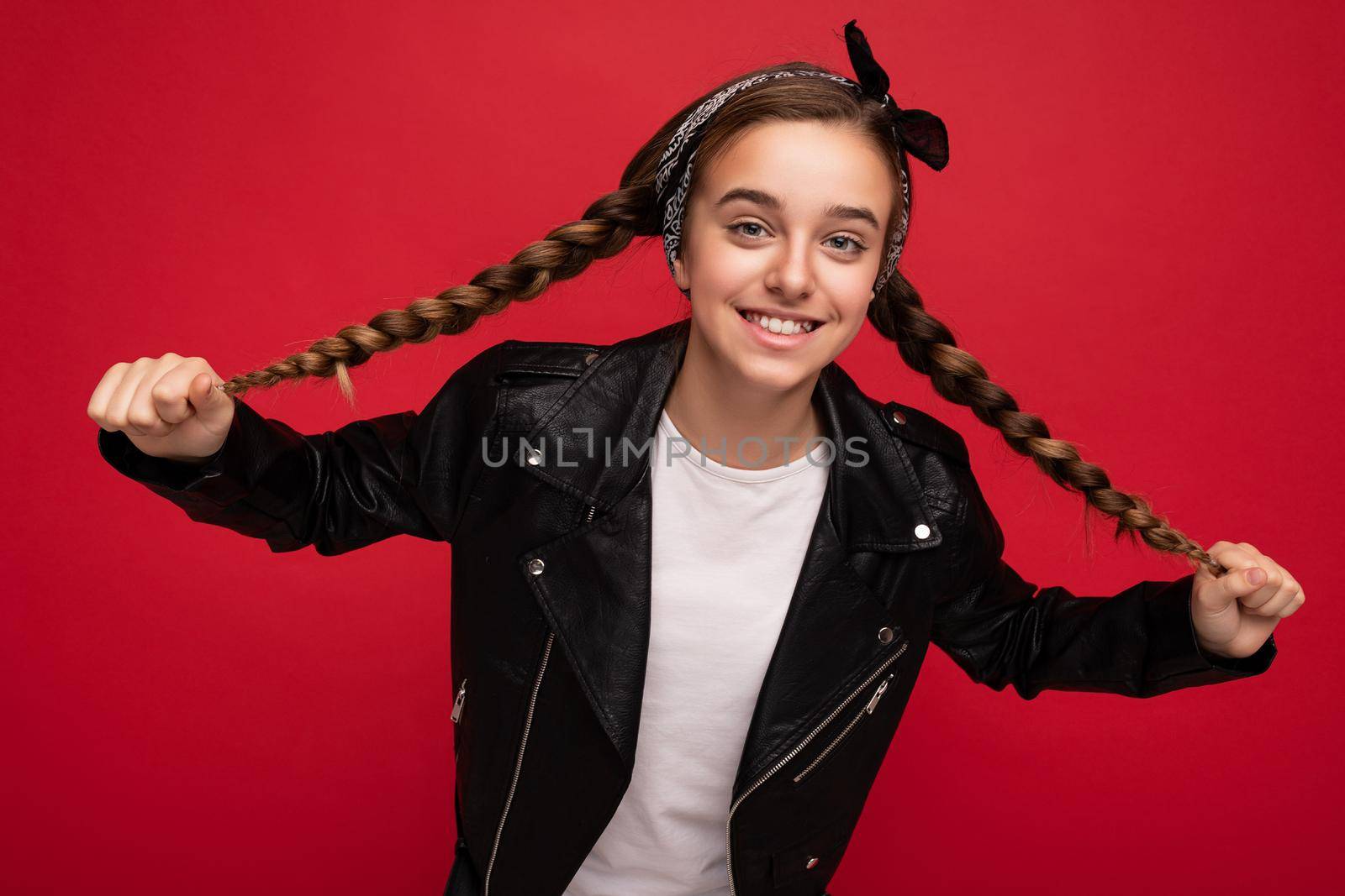 Portrait photo shot of pretty positive smiling brunette little girl with pigtails wearing stylish black leather jacket and white t-shirt for mockup standing isolated over red background wall looking at camera by TRMK