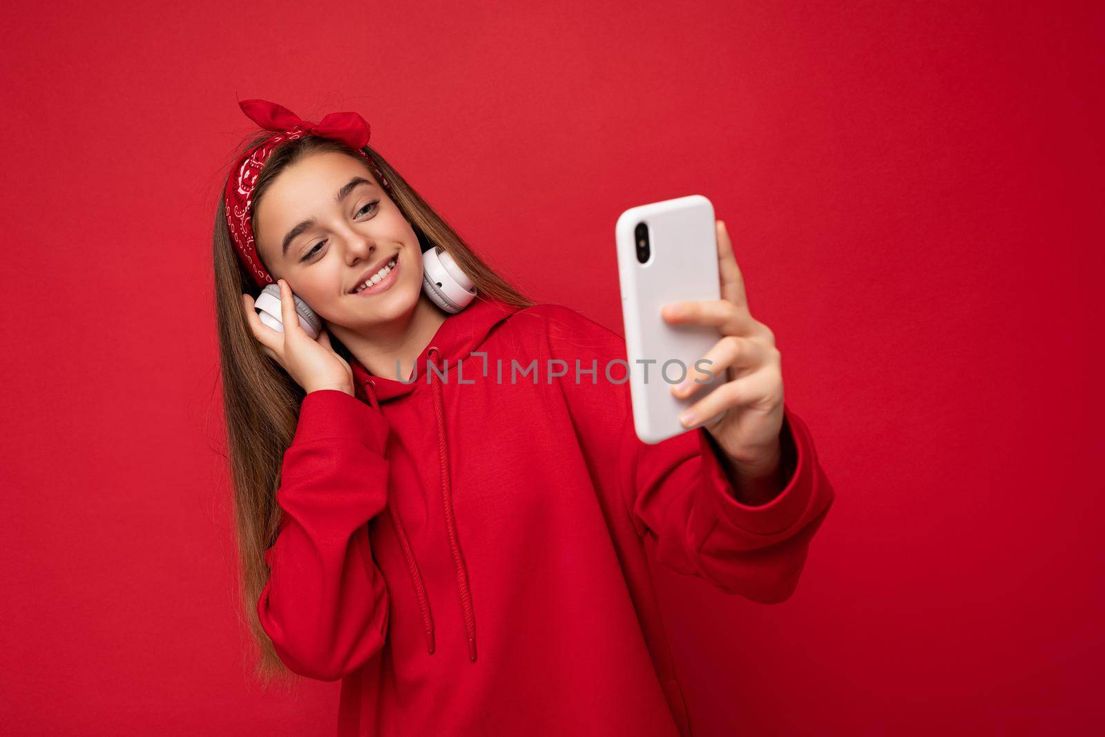Photo of beautiful happy smiling brunette girl wearing red hoodie isolated on red background holding and using smartphone wearing white wireless earphones listening to cool music looking at gadjet display by TRMK