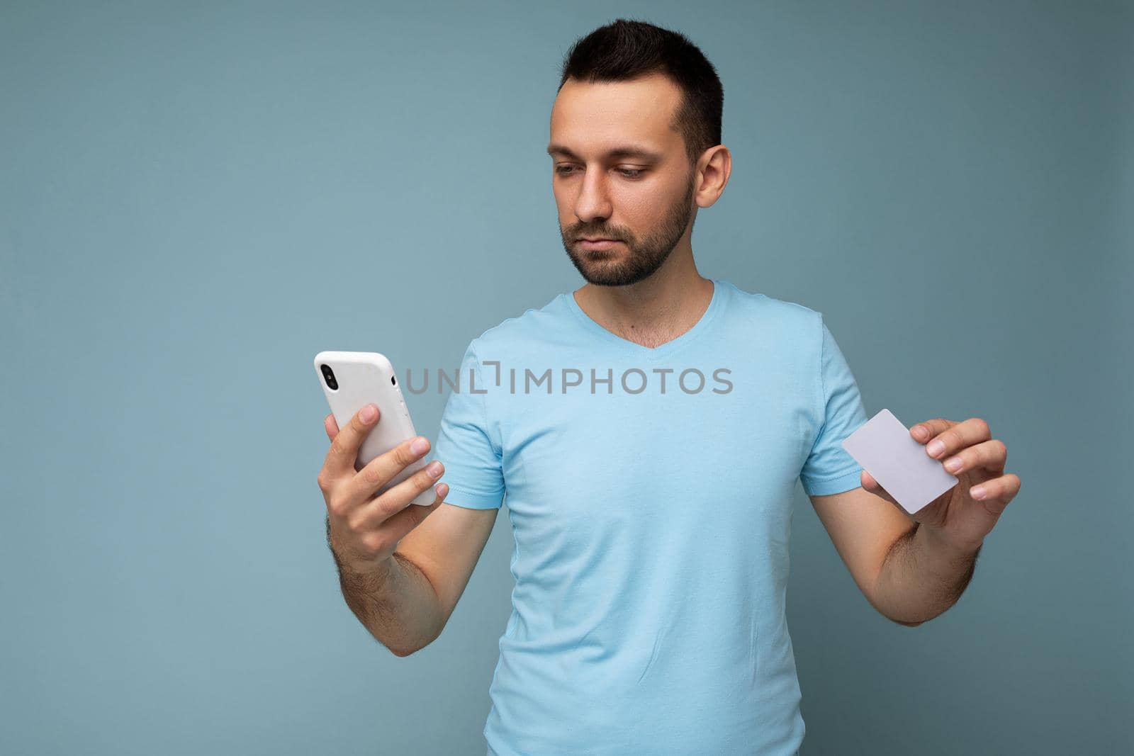Photo of handsome young unshaven brunet man with beard wearing everyday blue t-shirt isolated over blue background holding and using mobile phone and credit card making payment looking to the side.