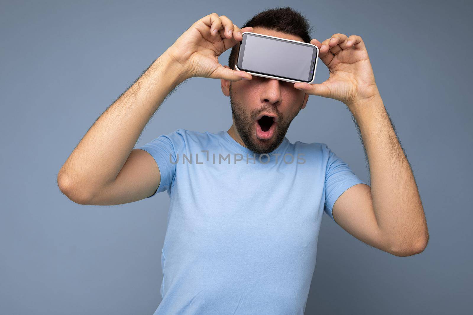 Positive amazed handsome young unshaven brunet man with beard wearing everyday blue t-shirt isolated over blue background holding and showing mobile phone with empty display for mockup.