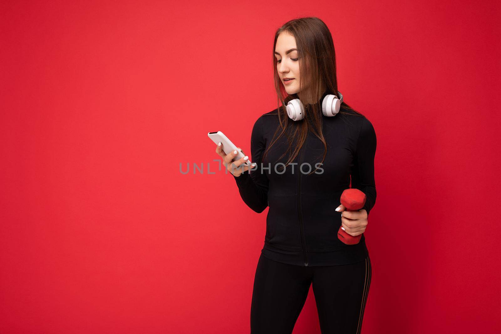 Photo of attractive positive young brunette woman wearing black sport clothes white headphones nad holding red dumbbell standing isolated over red background using mobile phone writing sms looking at gadjet display. copy space
