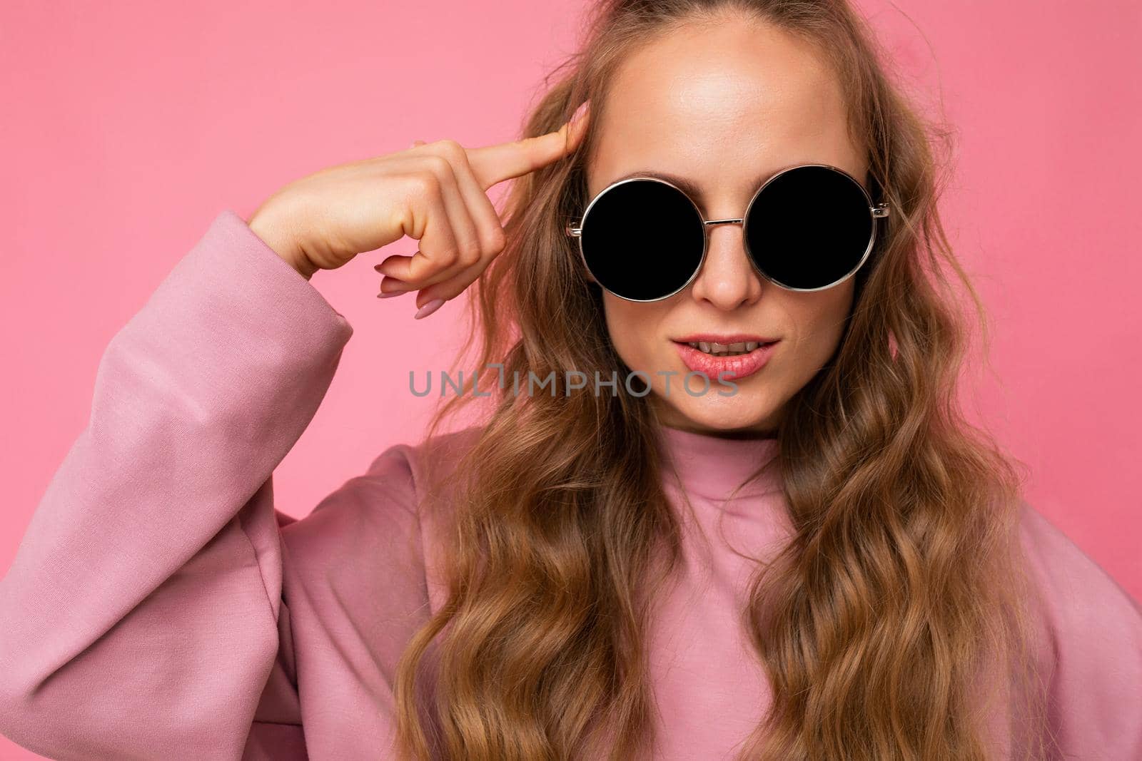 Closeup shot of beautiful serious young dark blonde curly woman isolated over pink background wall wearing casual pink sport clothes and stylish sunglasses looking at camera.