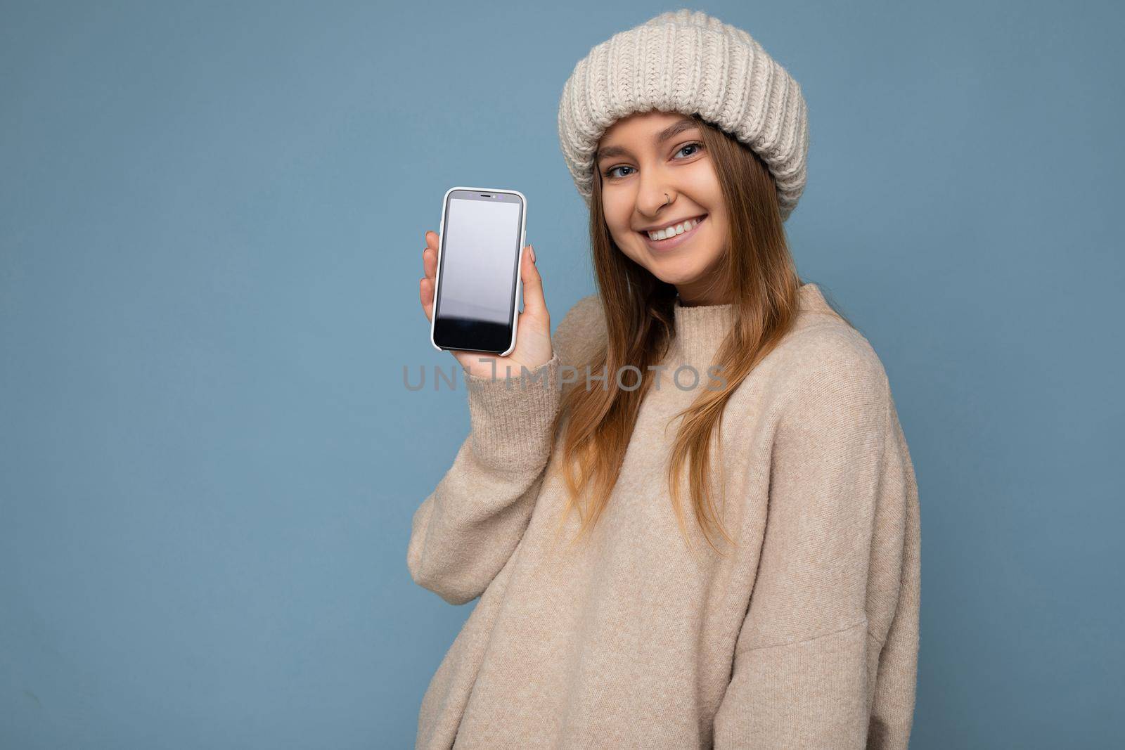 Photo of beautiful smiling positive good looking young female person wearing stylish beige sweater and beige knitted winter hat poising isolated over blue background with empty space holding in hand and showing mobile phone with empty screen for mockup looking at camera by TRMK