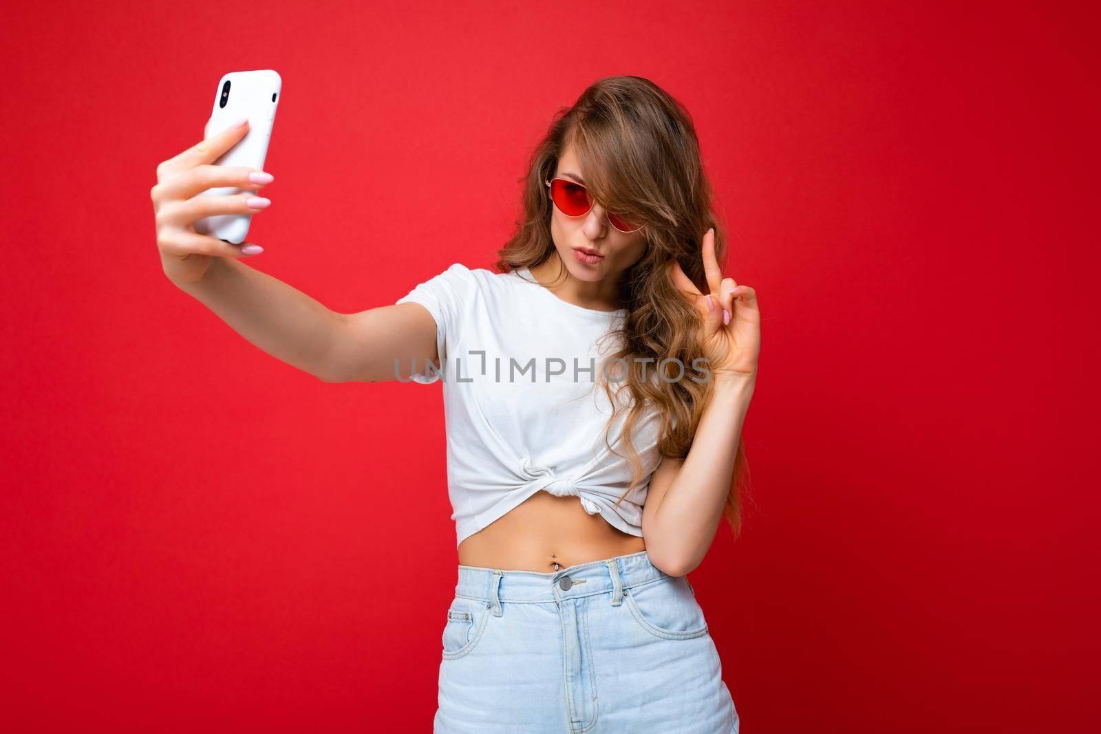 photo of amazing beautiful young blonde woman holding mobile phone taking selfie photo using smartphone camera wearing sunglasses everyday stylish outfit isolated over colorful wall background looking at device screen.