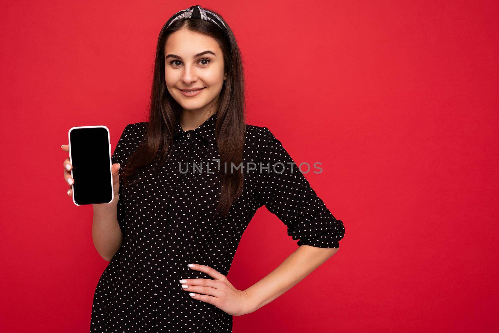 Photo of beautiful smiling girl good looking wearing casual stylish outfit standing isolated on background with copy space holding smartphone showing phone in hand with empty screen display for mockup looking at camera.