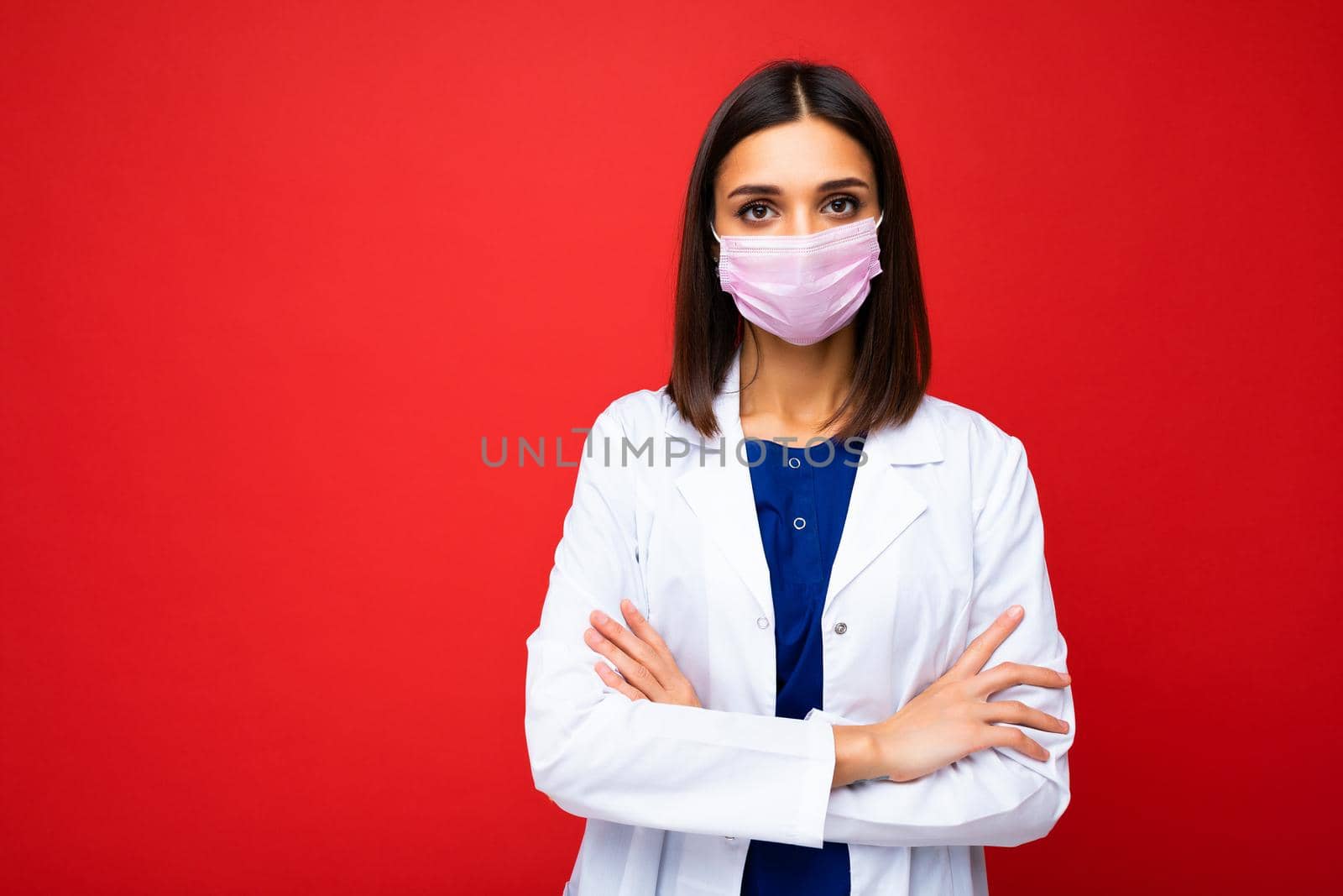 Young beautiful brunette woman in virus protective mask on face against coronavirus and white medical coat isolated on the background by TRMK